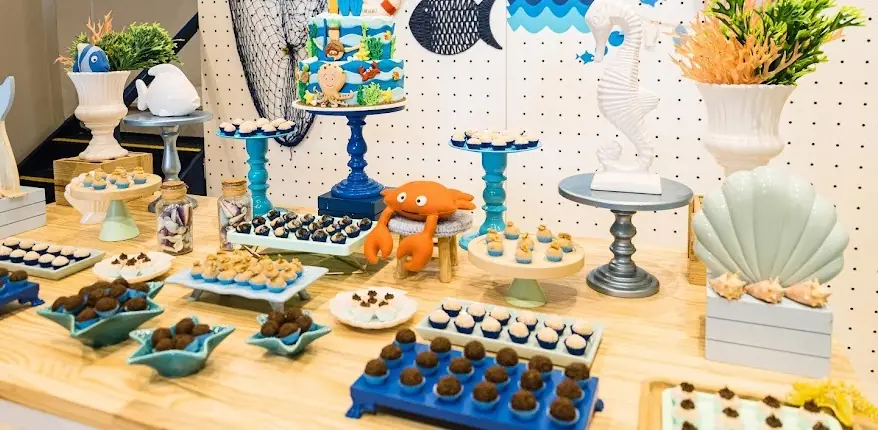 Mesa decorada com tema fundo do mar: bolo de andares, docinhos em bandejas azuis, boleiras e personagens como caranguejo e cavalo-marinho